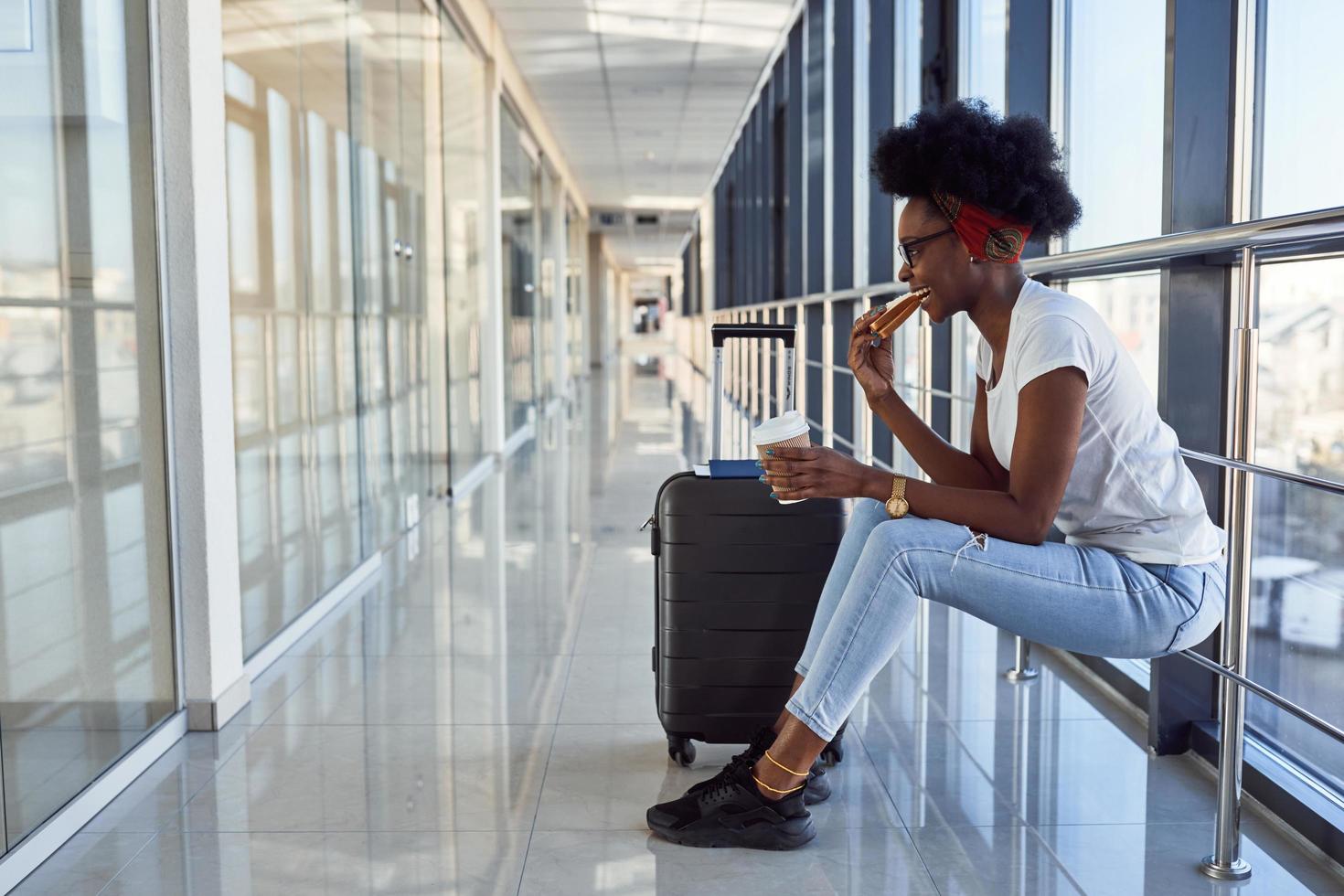 Young african american female passanger in casual clothes is in airport with baggage eating some food photo