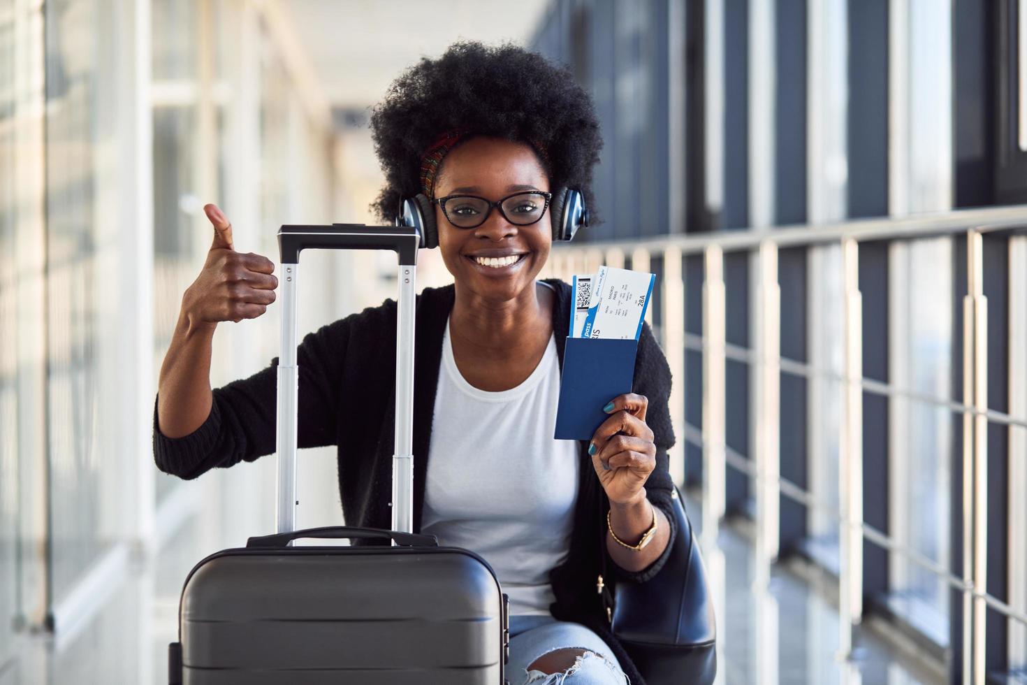 Young african american female passanger in casual clothes and headphones is in airport with baggage photo