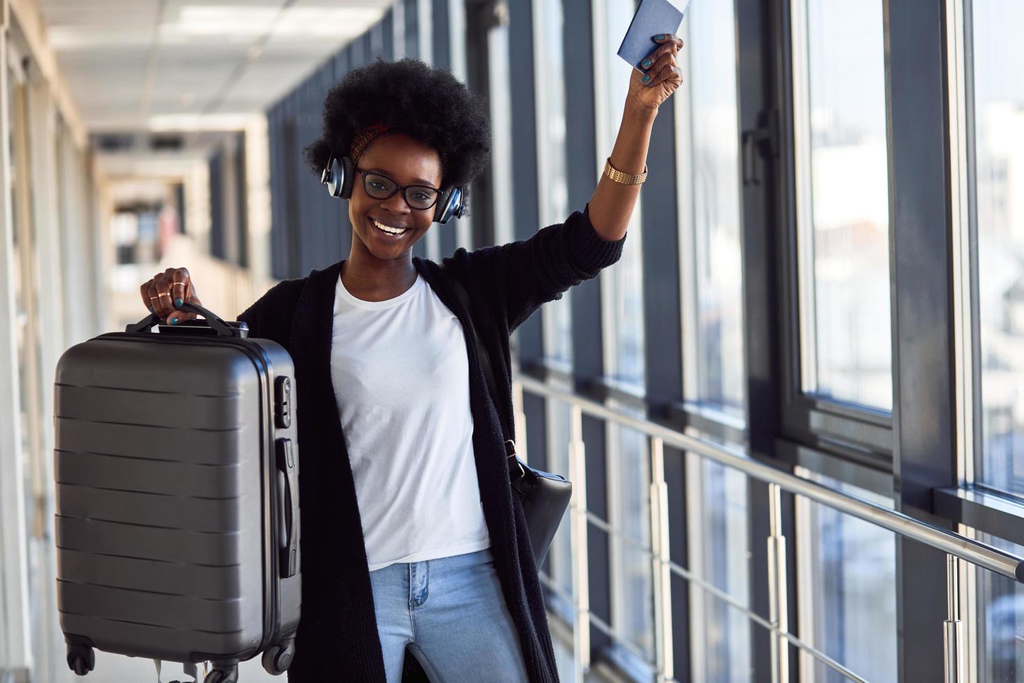 una joven pasajera afroamericana con ropa informal y auriculares está en el aeropuerto con equipaje foto