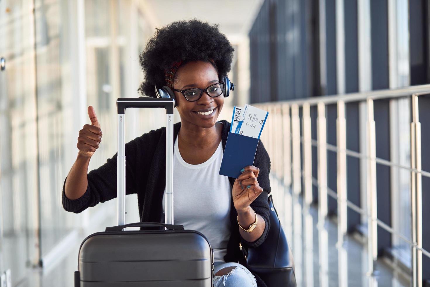 Young african american female passanger in casual clothes and headphones is in airport with baggage photo