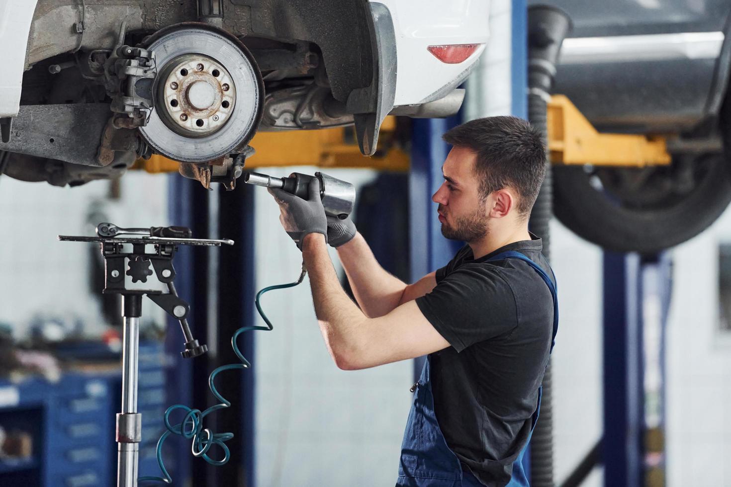 Using special tool. Man in work uniform repairs white automobile indoors. Conception of car service photo