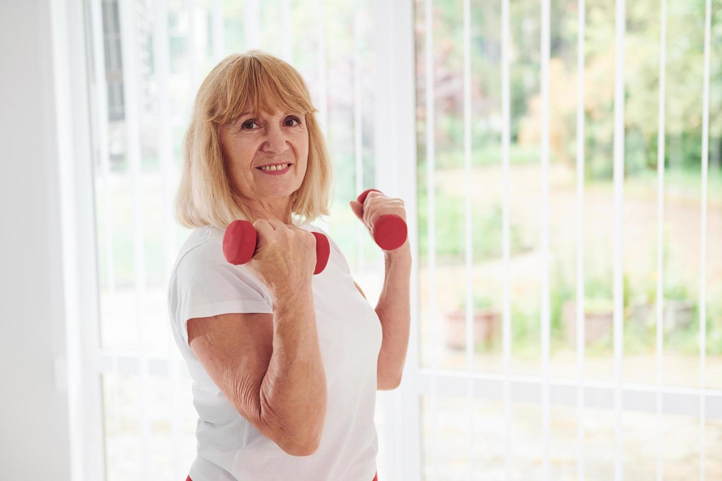 Positive senior woman in sportive clothes indoors at daytime doing exercises with dumbbells photo