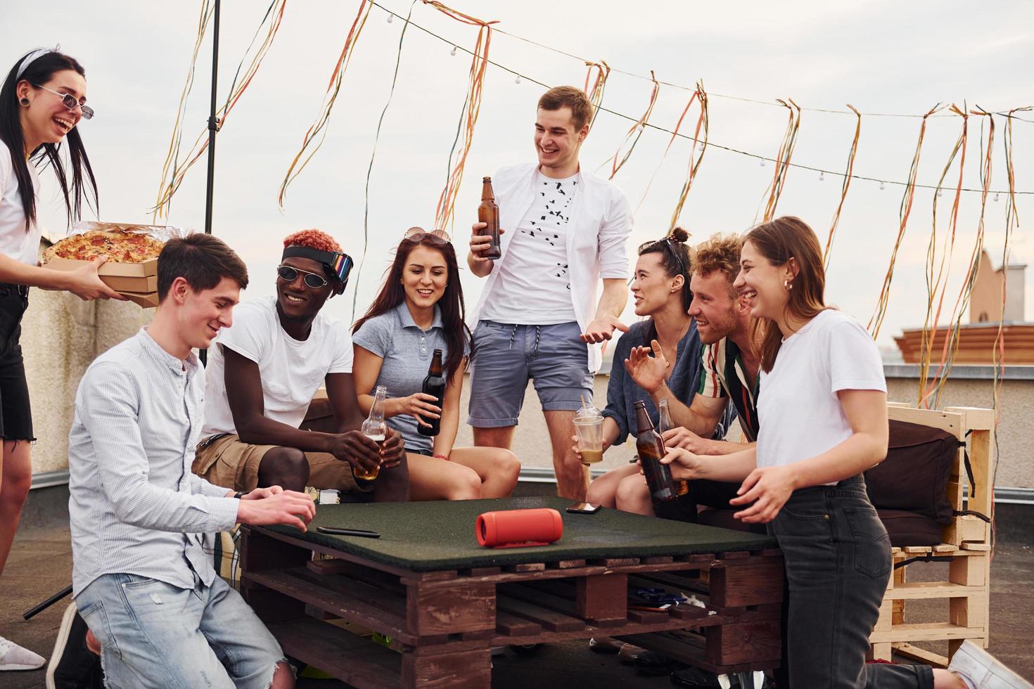 con deliciosa pizza. un grupo de jóvenes con ropa informal tienen una fiesta en la azotea juntos durante el día foto