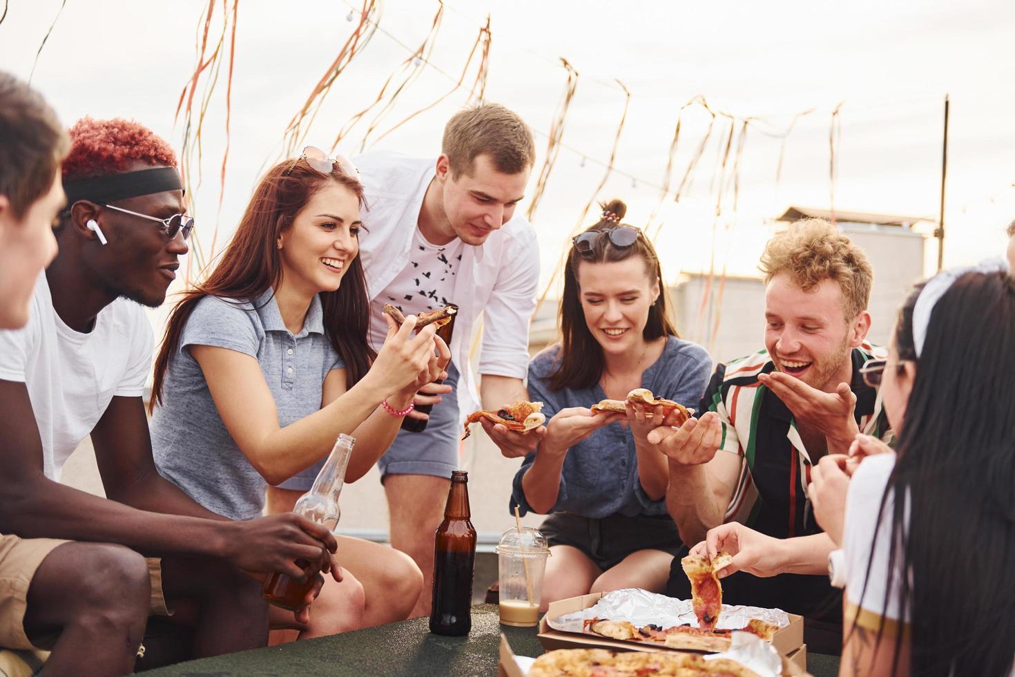 con deliciosa pizza. un grupo de jóvenes con ropa informal tienen una fiesta en la azotea juntos durante el día foto