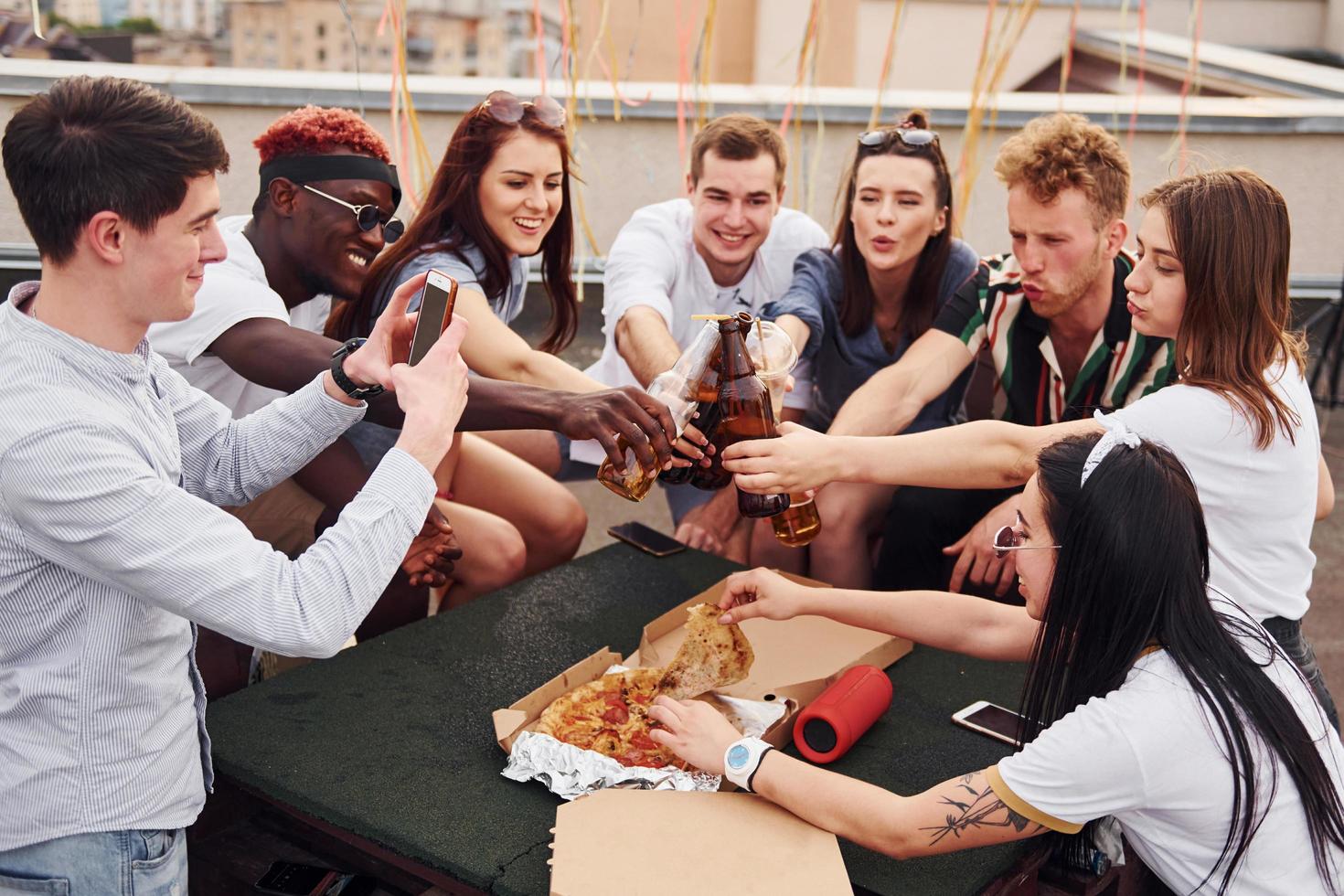 Doing cheers by bottles with beer. Group of young people in casual clothes have a party at rooftop together at daytime photo