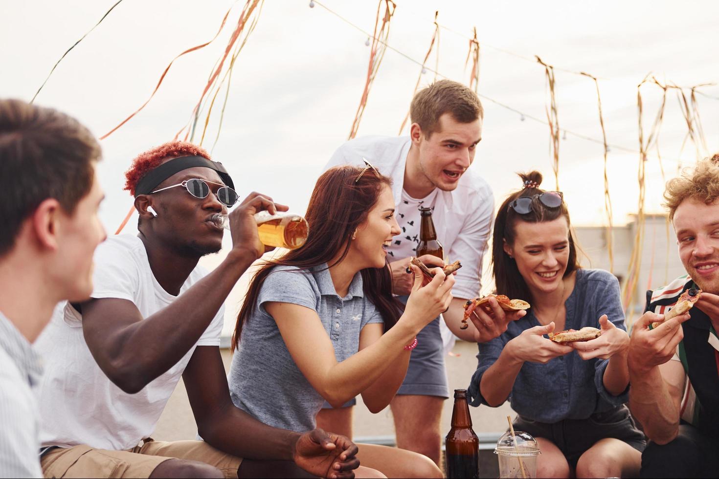 con deliciosa pizza. un grupo de jóvenes con ropa informal tienen una fiesta en la azotea juntos durante el día foto