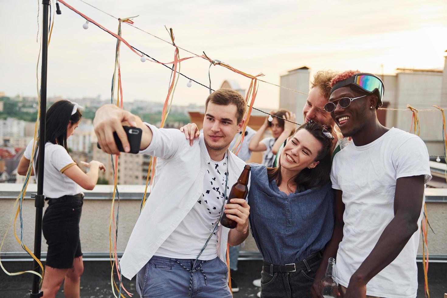 Group of young people in casual clothes have a party at rooftop together at daytime and doing selfie by phone photo