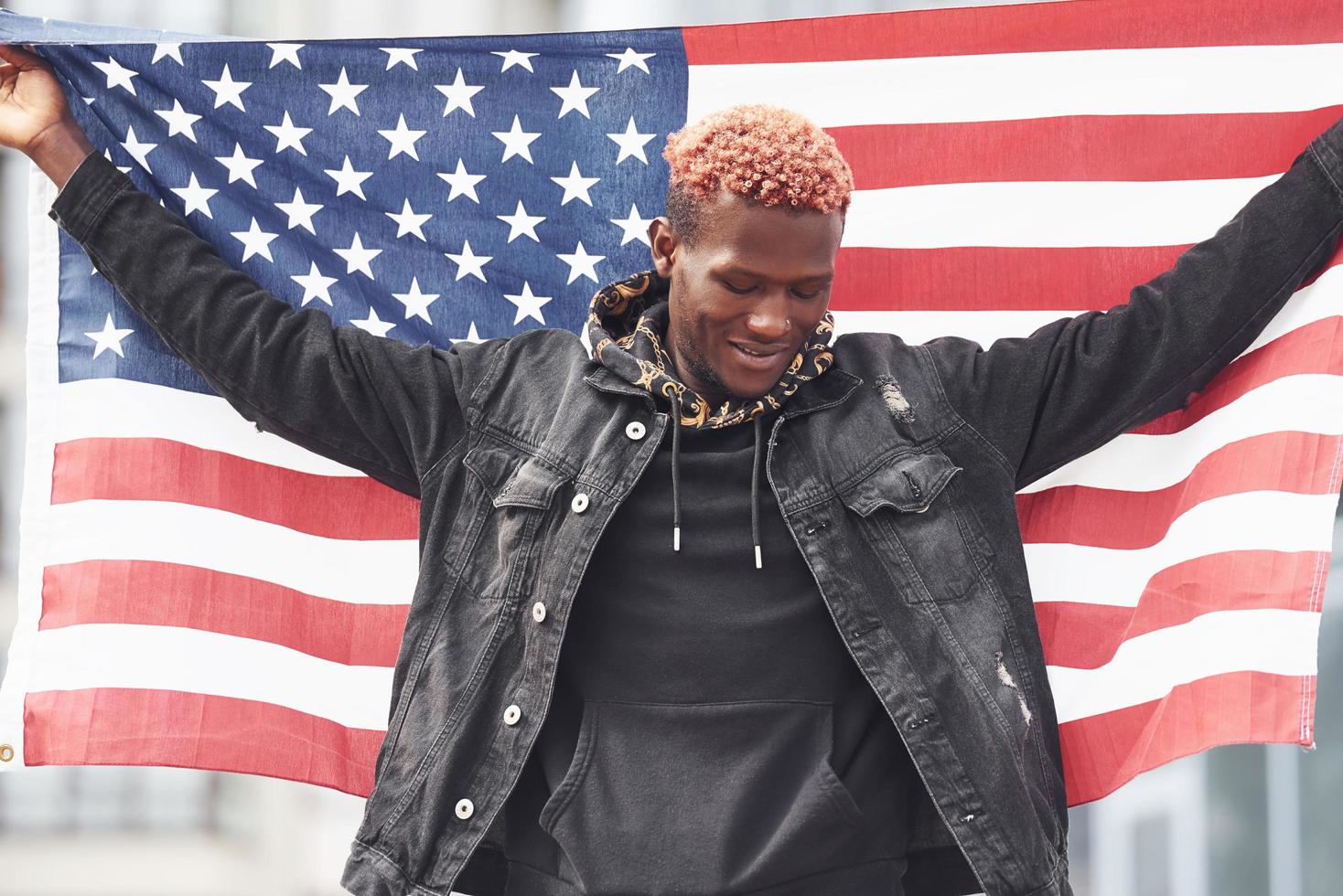 Patriot holding USA flag. Conception of pride and freedom. Young african american man in black jacket outdoors in the city standing against modern business building photo