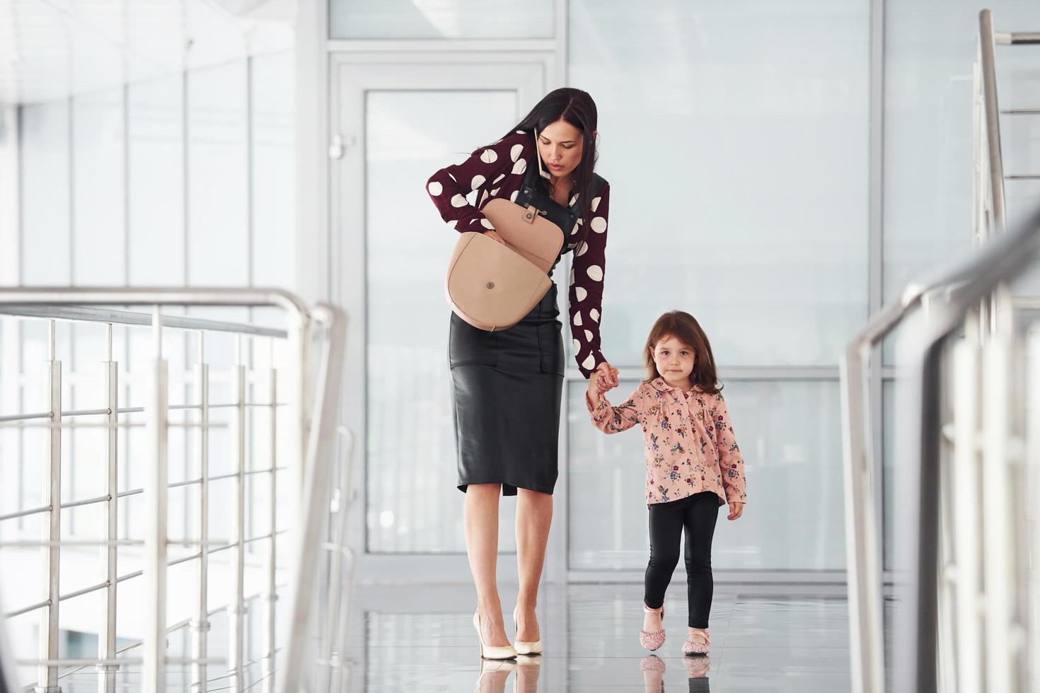 Young mother with her daughter walking together indoors in the office or airport. Having vacation photo
