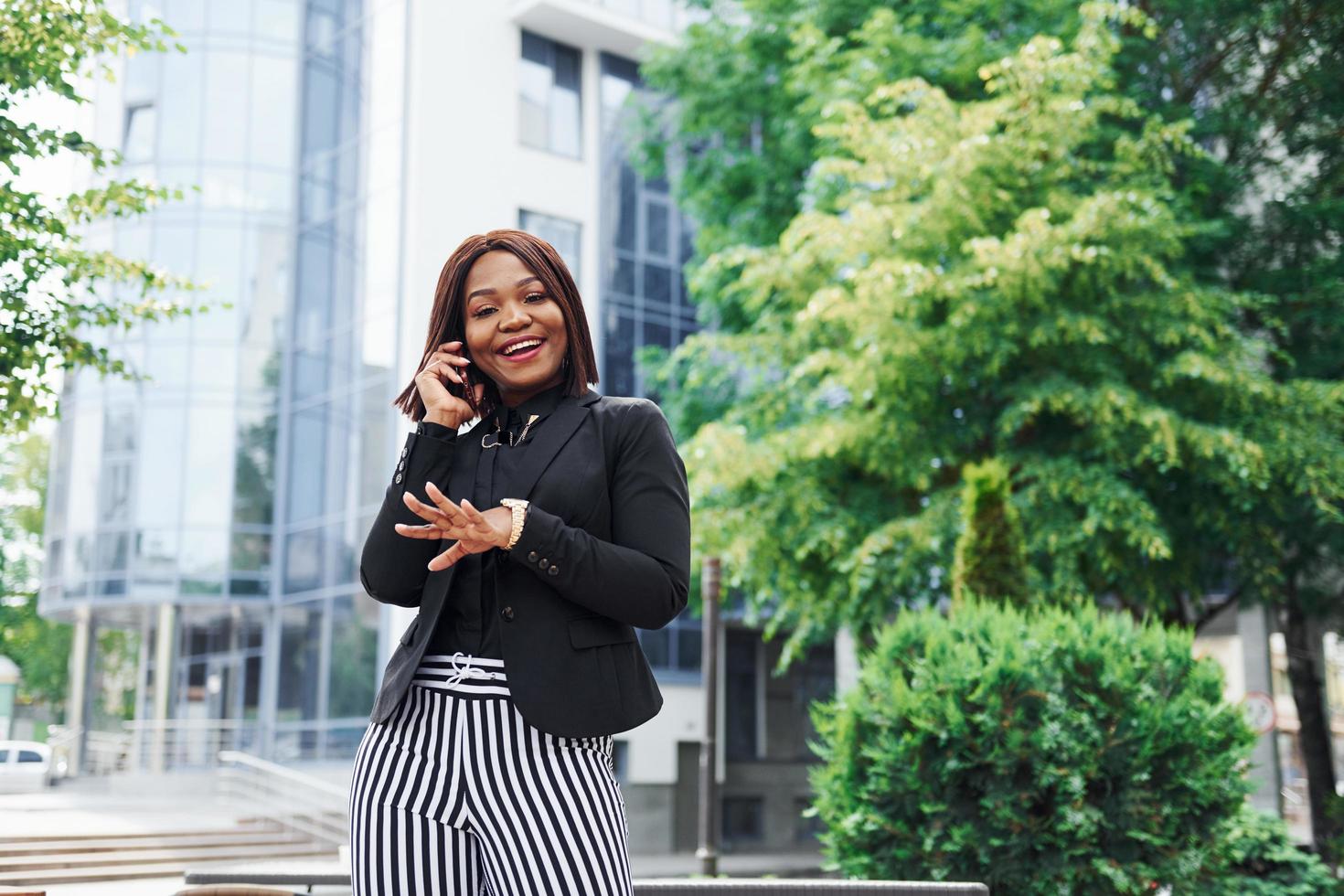 Talking by phone. Young afro american woman in fashionable clothes outdoors in the city near green trees and against business building photo