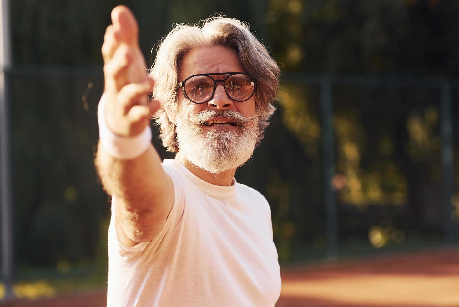 hombre mayor con estilo en anteojos, camisa blanca y pantalones cortos deportivos negros en la cancha de tenis foto