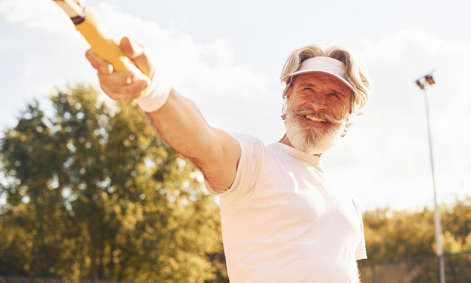 hermosa luz del sol. hombre mayor con estilo en camisa blanca y pantalones cortos deportivos negros en la cancha de tenis foto