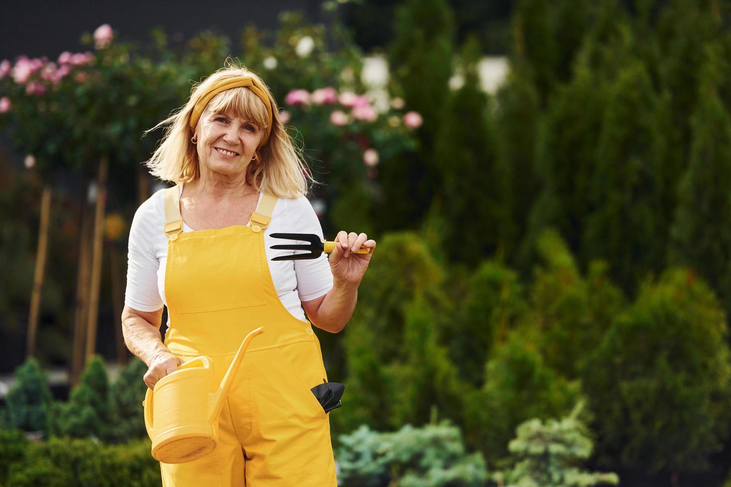 Holding yellow watering can. Senior woman is in the garden at daytime. Conception of plants and seasons photo