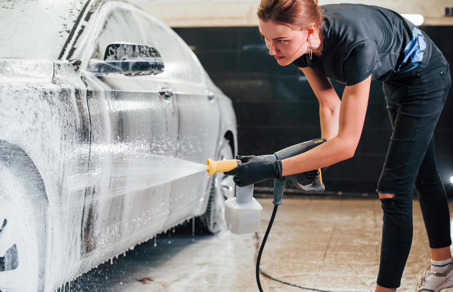 utilizando agua a alta presión. el automóvil negro moderno es limpiado por una mujer dentro de la estación de lavado de autos foto