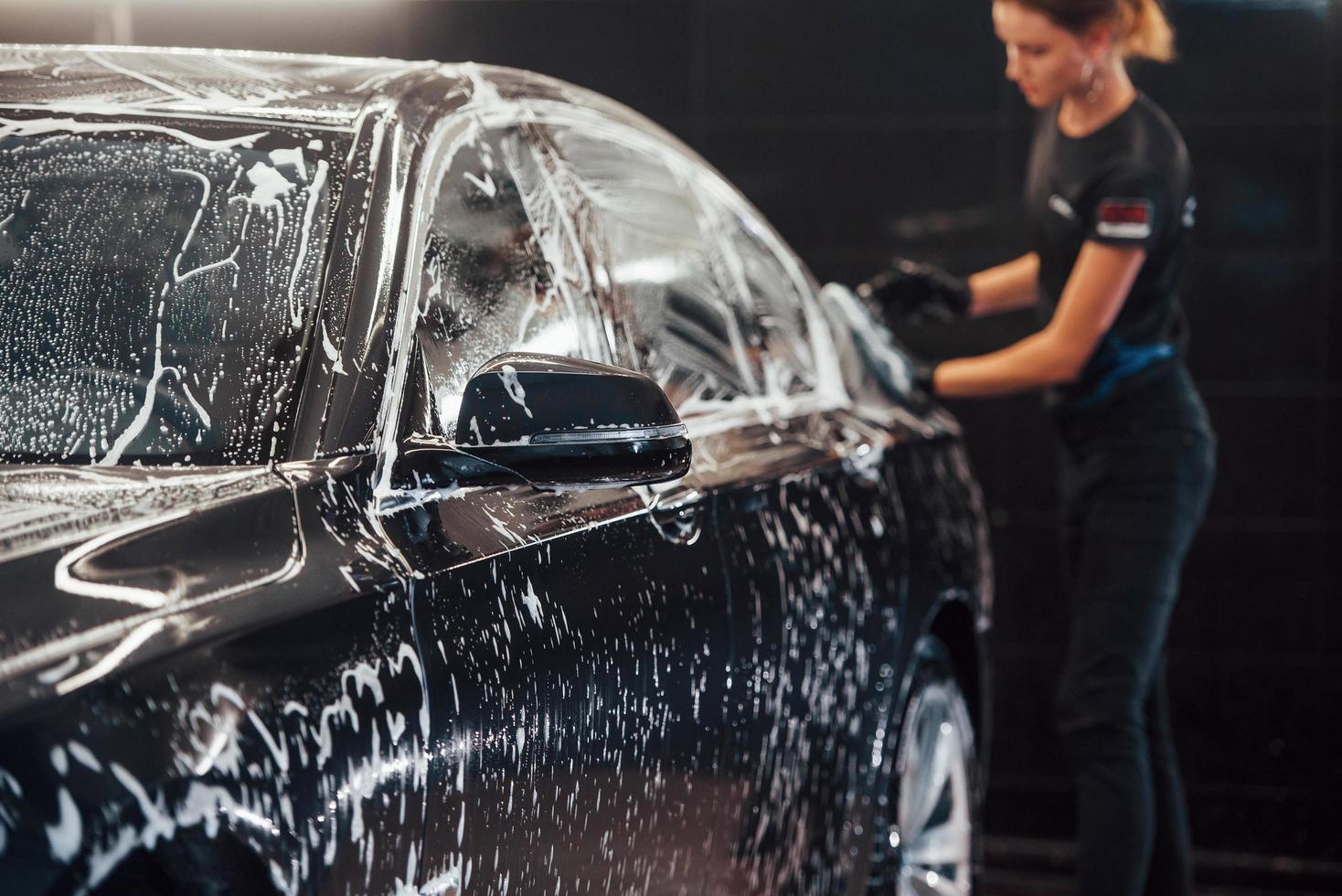 Wipes vehicle that is in white soap. Modern black automobile get cleaned by woman inside of car wash station photo