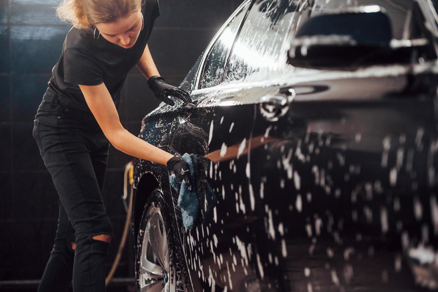 Wipes vehicle that is in white soap. Modern black automobile get cleaned by woman inside of car wash station photo