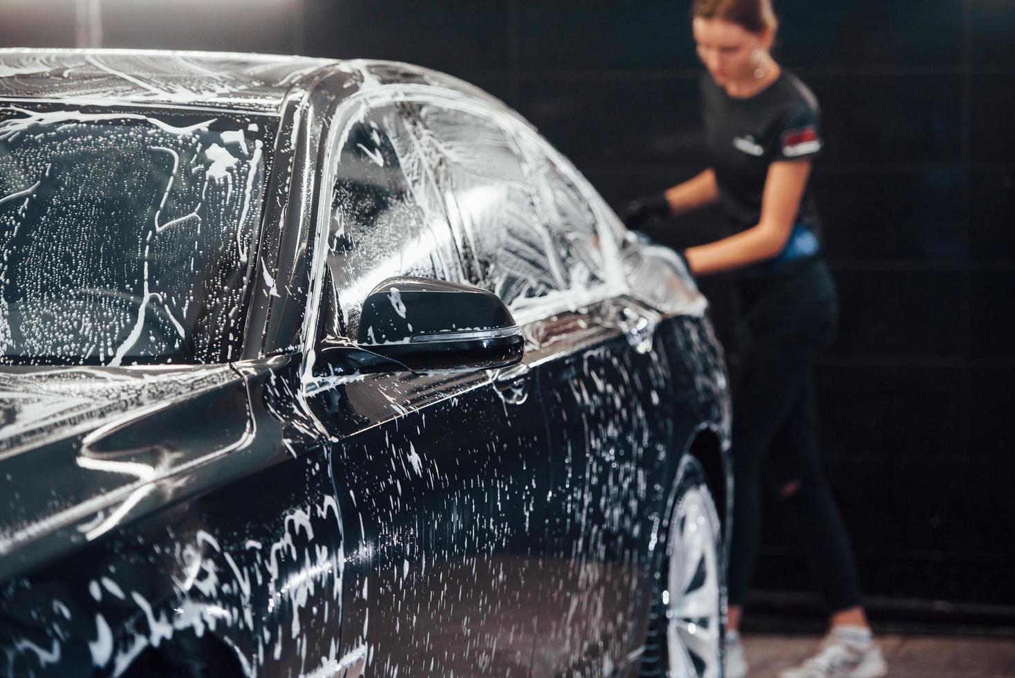 Wipes vehicle that is in white soap. Modern black automobile get cleaned by woman inside of car wash station photo