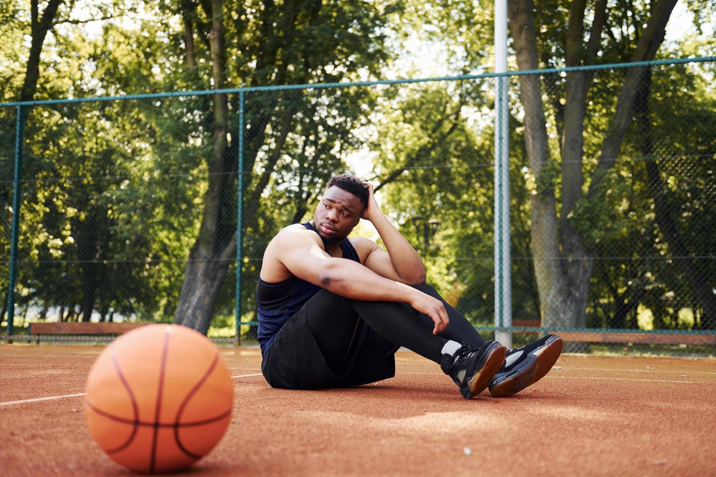 Tired african american man sits on the ground with ball on the court outdoors. Takes a break photo