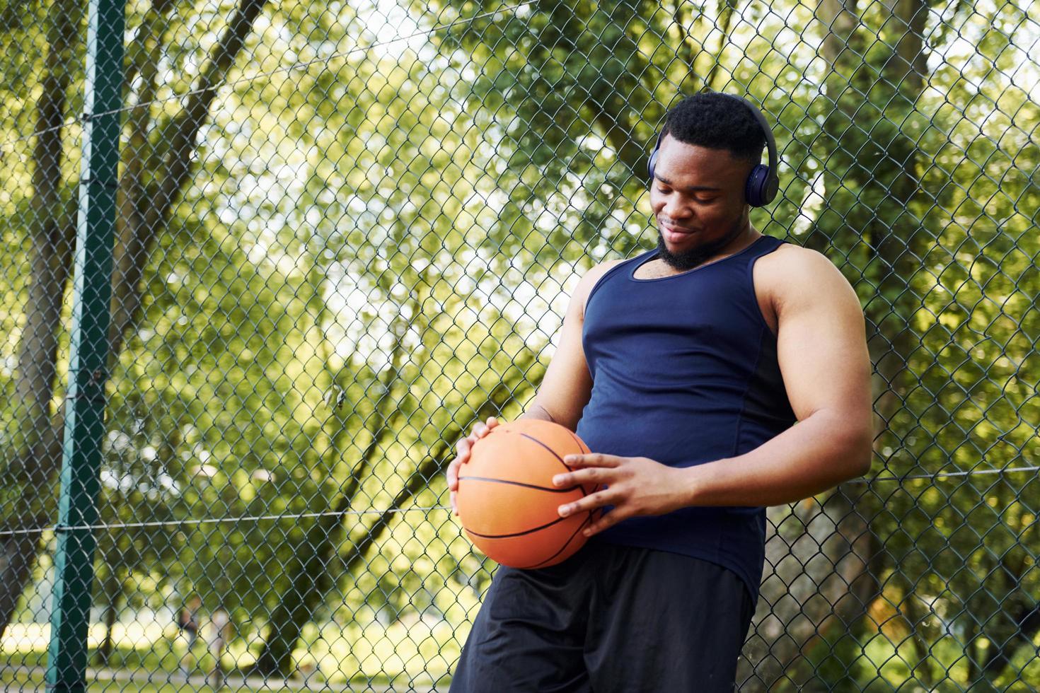 African american man with wireless headphones takes a break and leaning on the metal mesh with ball on the court outdoors photo