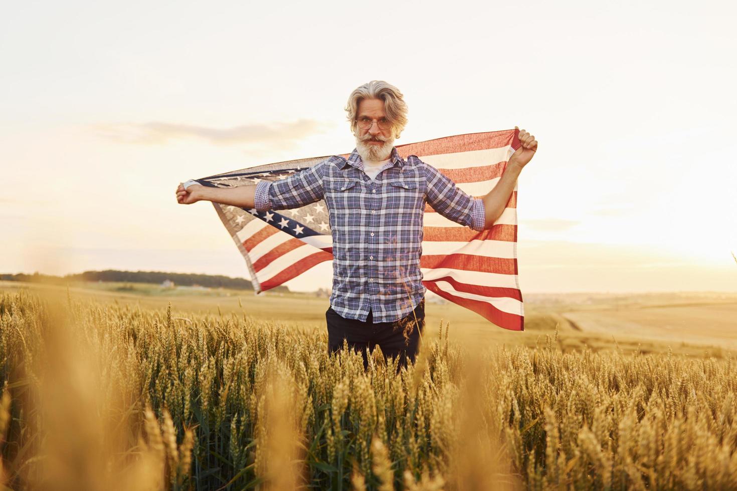 Holding USA flag in hands. Patriotic senior stylish man with grey hair and beard on the agricultural field photo