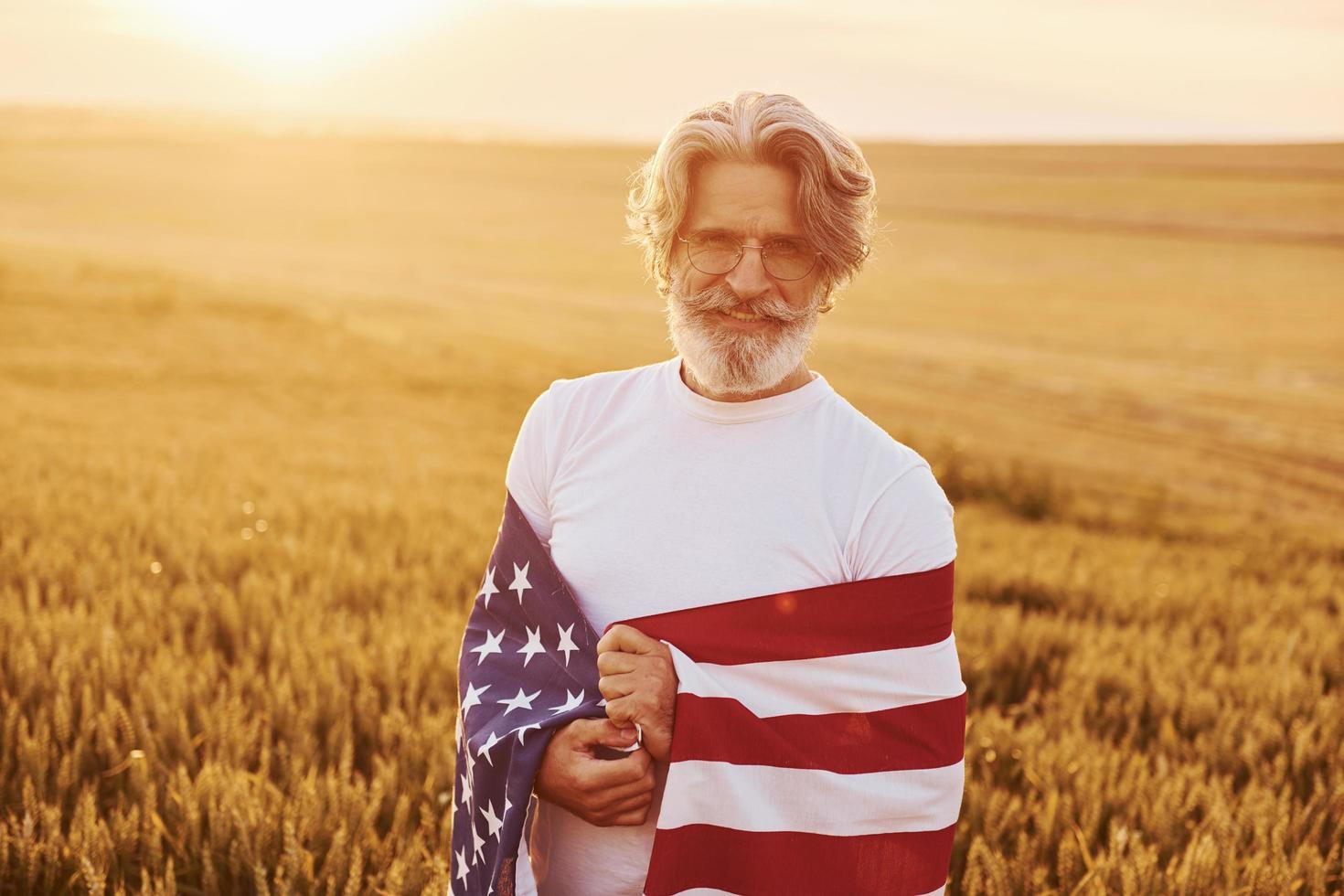 Holding USA flag in hands. Patriotic senior stylish man with grey hair and beard on the agricultural field photo