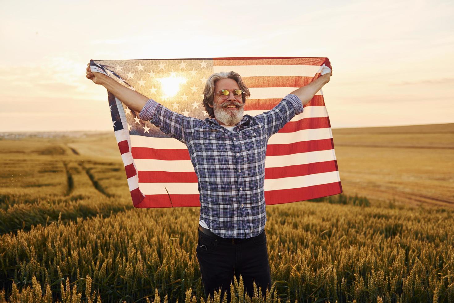 Holding USA flag in hands. Patriotic senior stylish man with grey hair and beard on the agricultural field photo