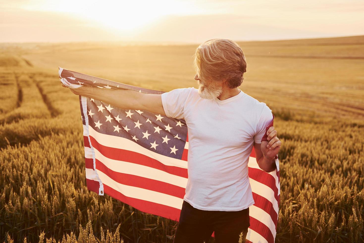 Holding USA flag in hands. Patriotic senior stylish man with grey hair and beard on the agricultural field photo