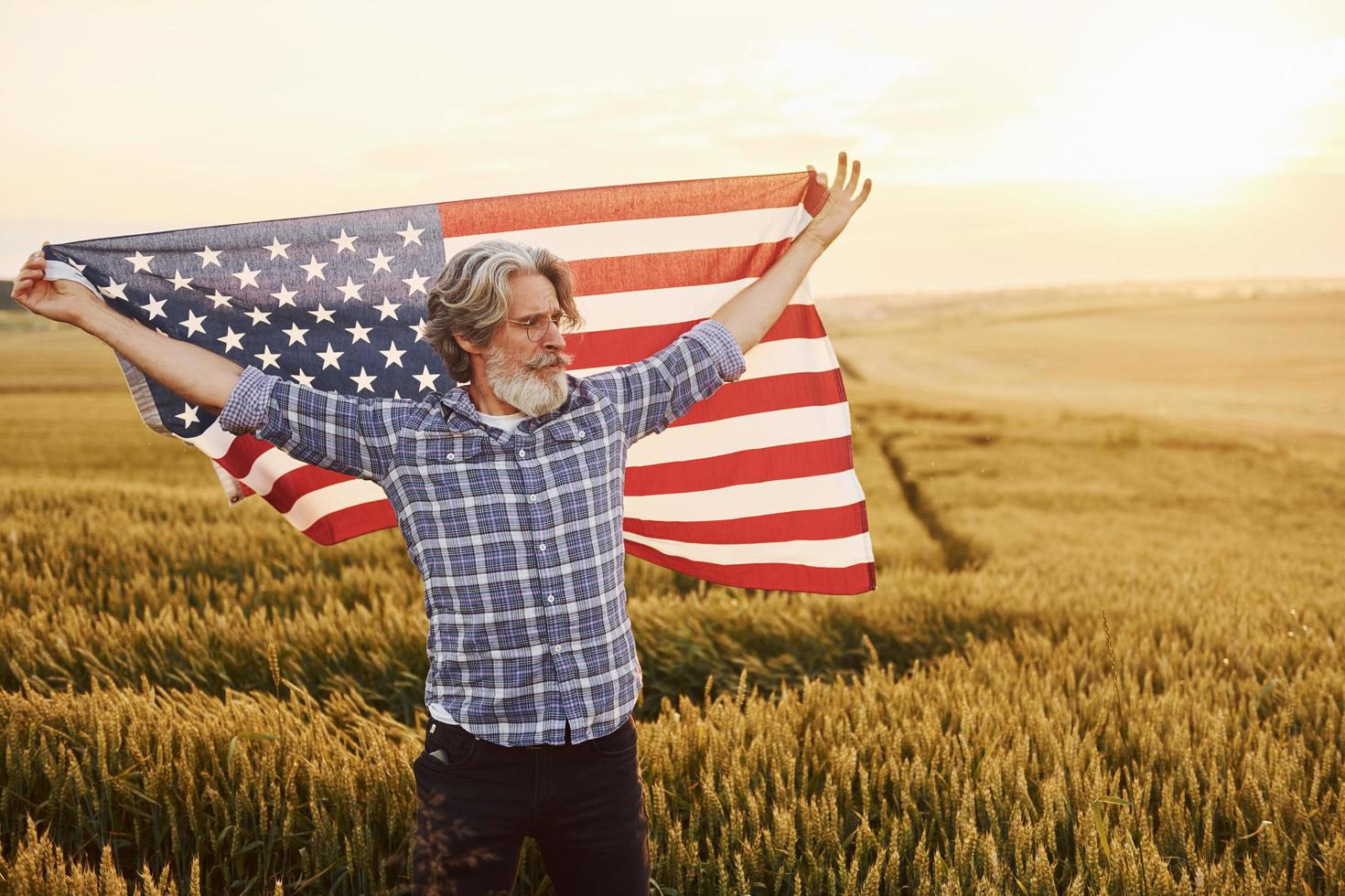 Holding USA flag in hands. Patriotic senior stylish man with grey hair and beard on the agricultural field photo