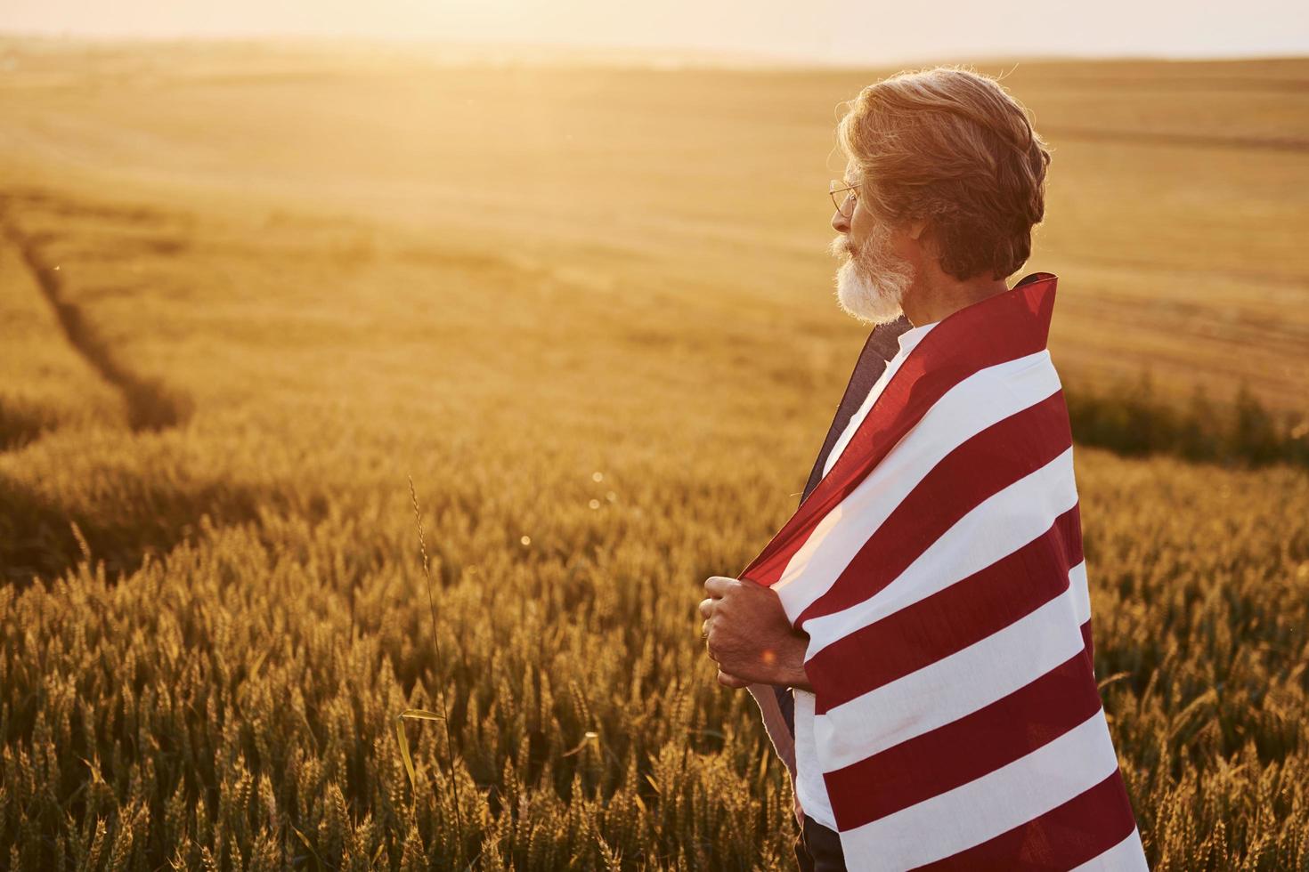View from behind. Holding USA flag in hands. Patriotic senior stylish man with grey hair and beard on the agricultural field photo
