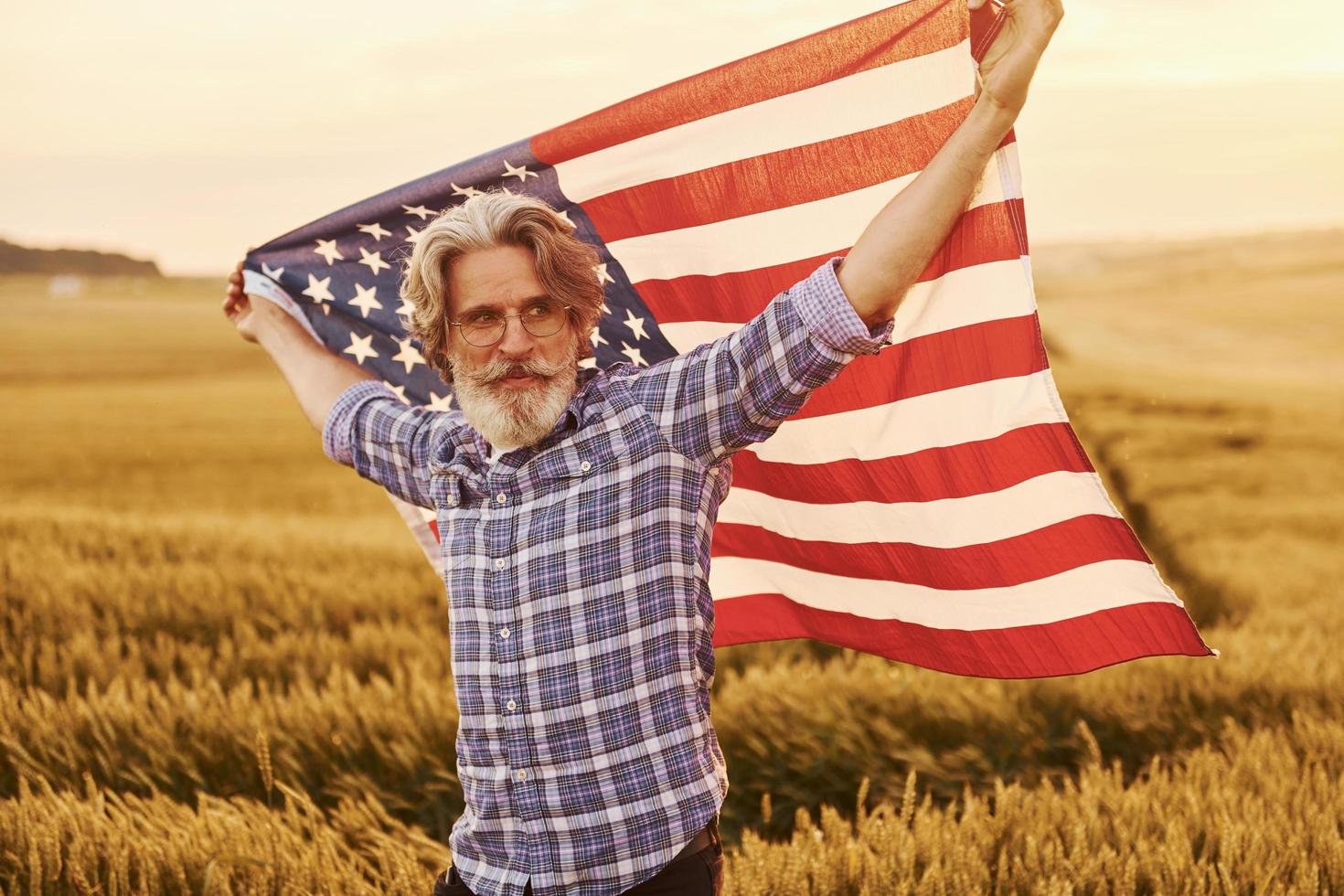 Holding USA flag in hands. Patriotic senior stylish man with grey hair and beard on the agricultural field photo