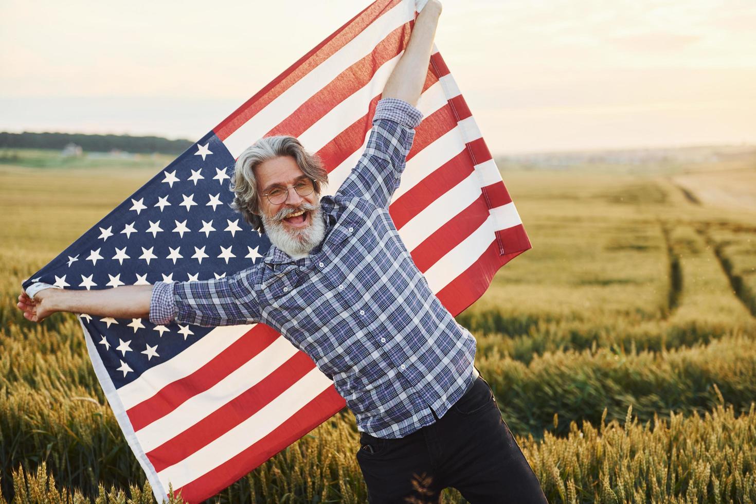 Holding USA flag in hands. Patriotic senior stylish man with grey hair and beard on the agricultural field photo