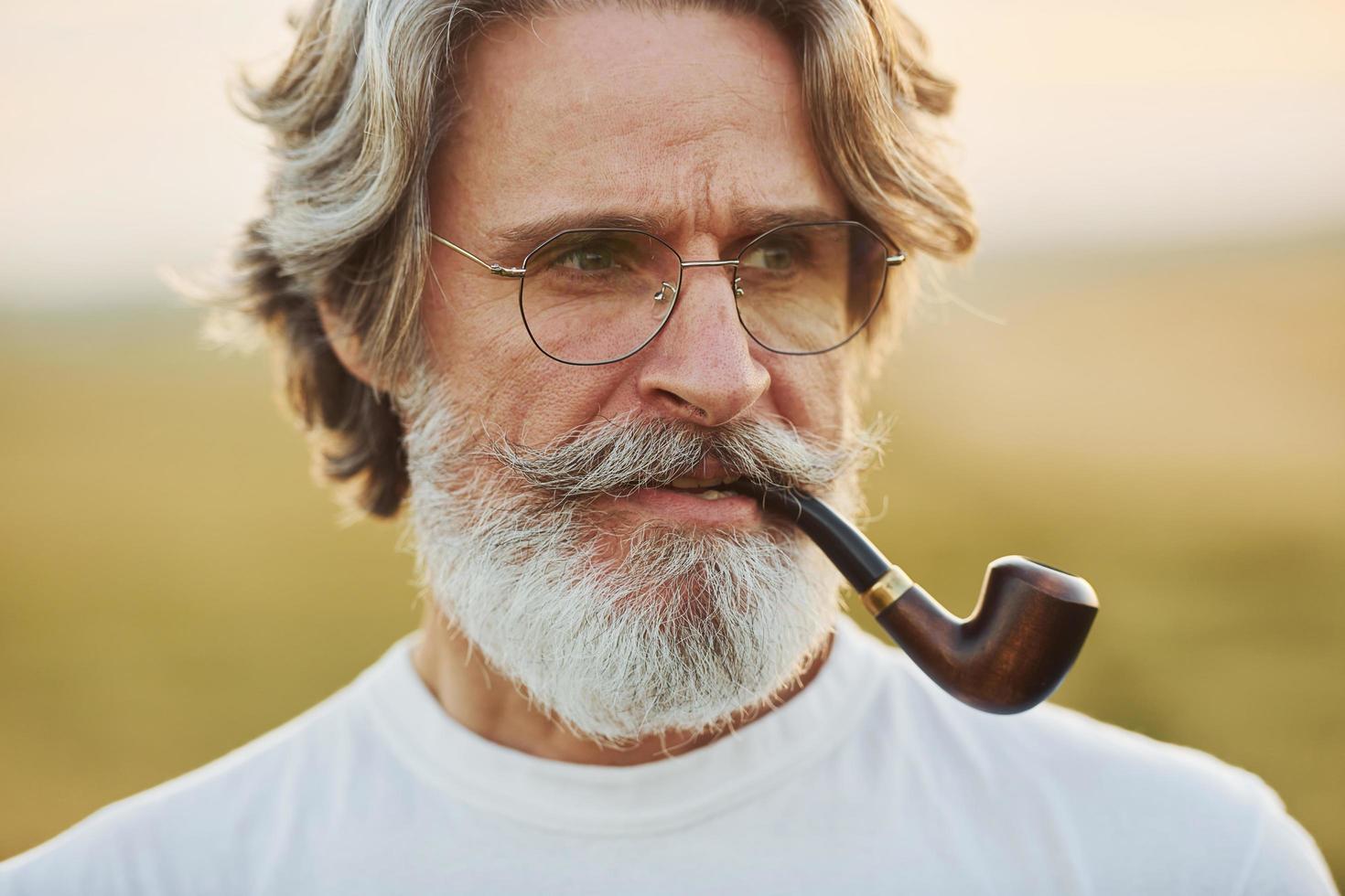 Portrait of senior stylish man with grey hair and beard that standing outdoors on field at sunny day and smoking photo