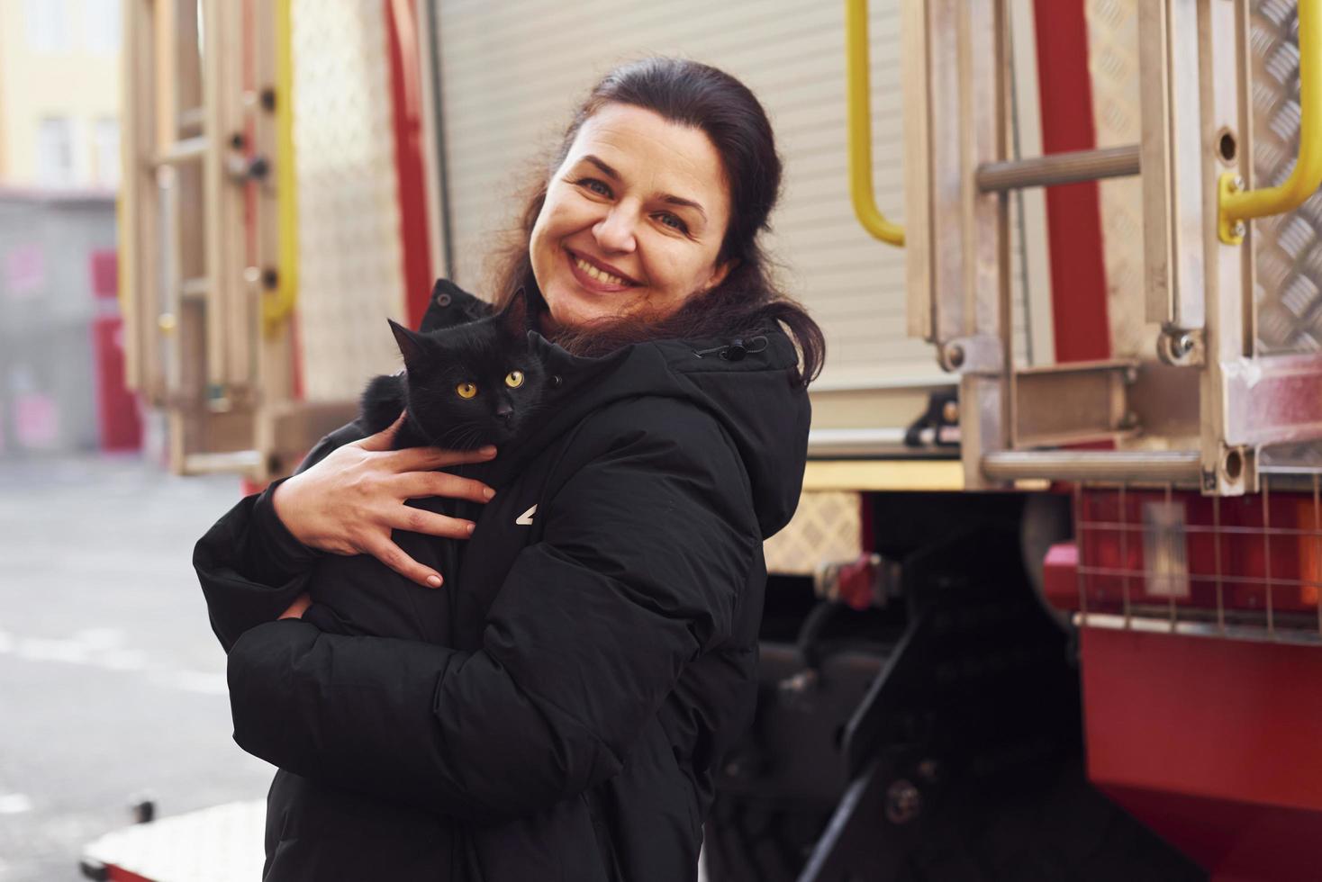 Happy woman holds her black cat in hands ner fire truck outdoors photo