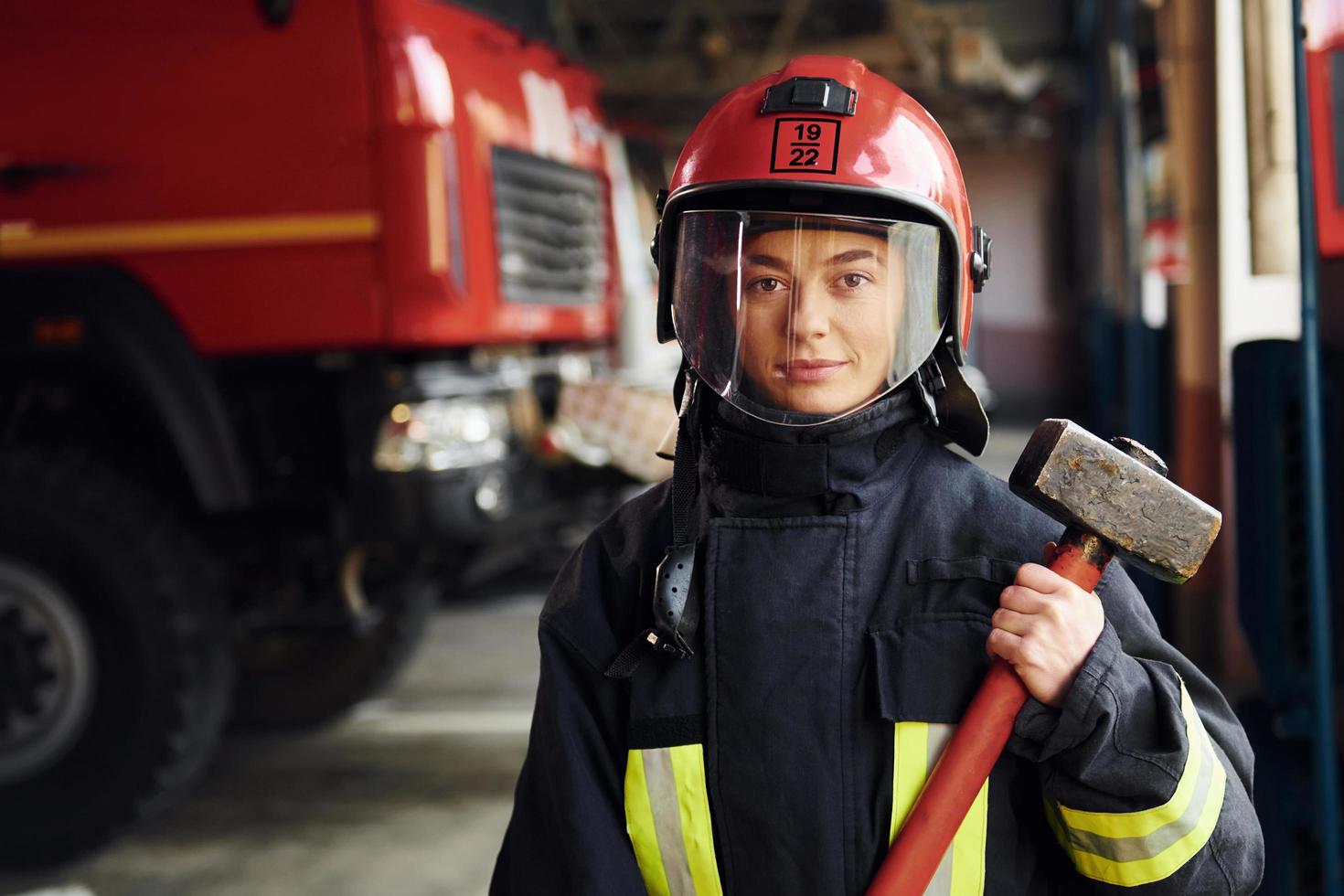 con martillo en las manos. bombero femenino en uniforme protector de pie cerca de camión foto