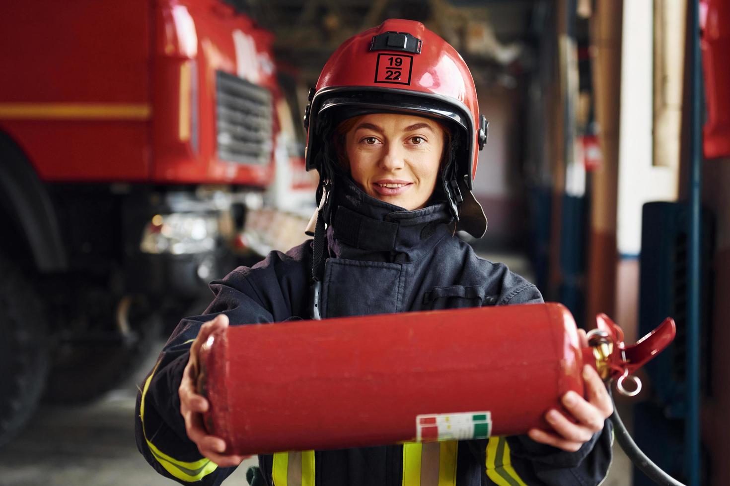 Holds extinguisher in hands. Female firefighter in protective uniform standing near truck photo