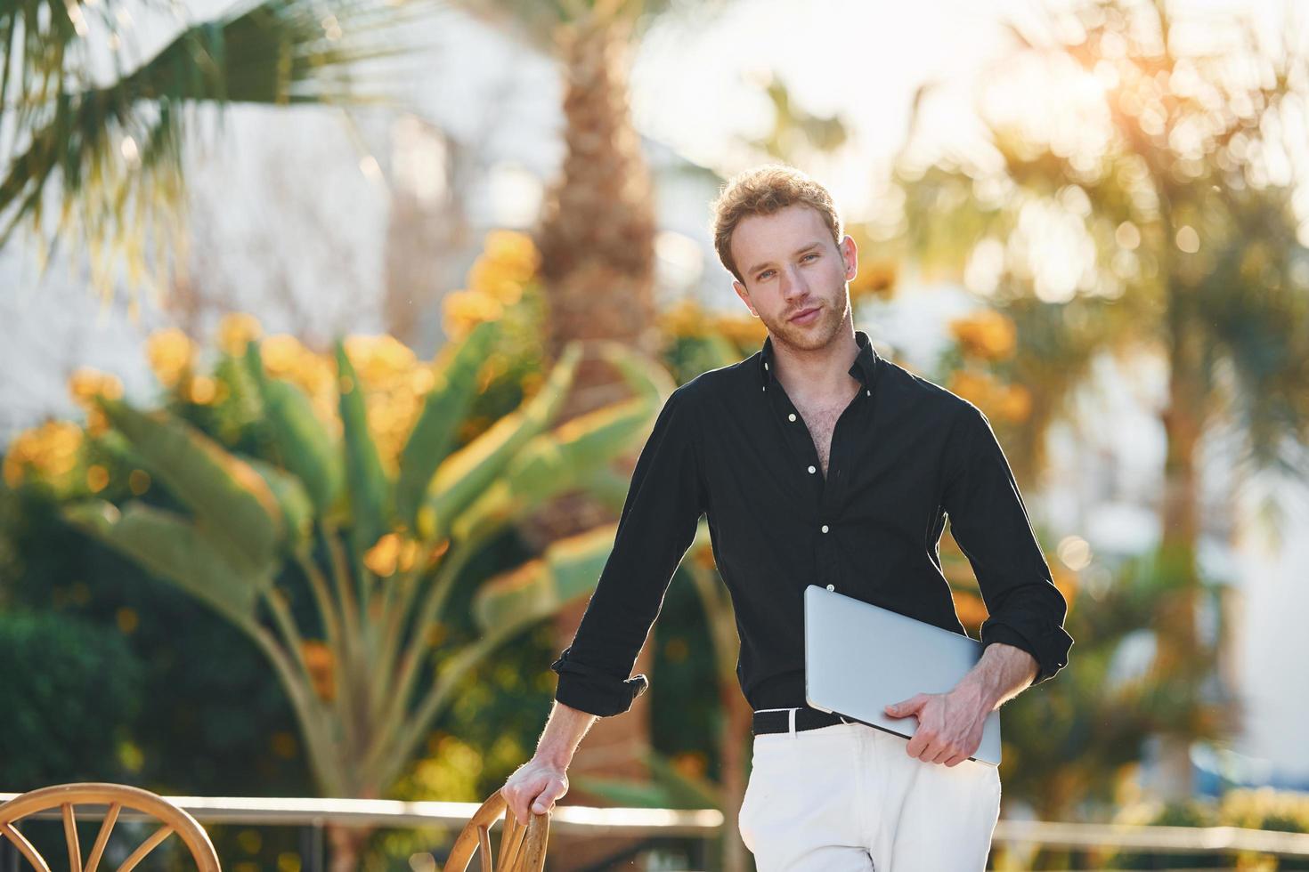 Holds laptop. Young man is outdoors at sunny daytime. Concept of vacation photo