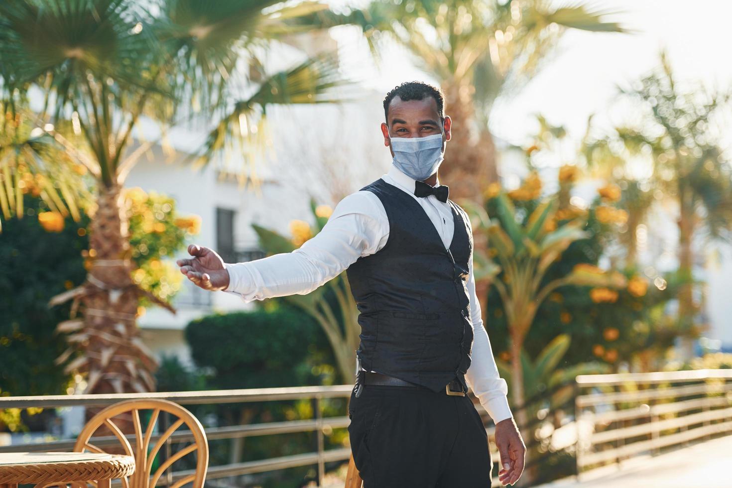 en máscara protectora. camarero negro con ropa formal está en su trabajo al aire libre durante el día soleado foto