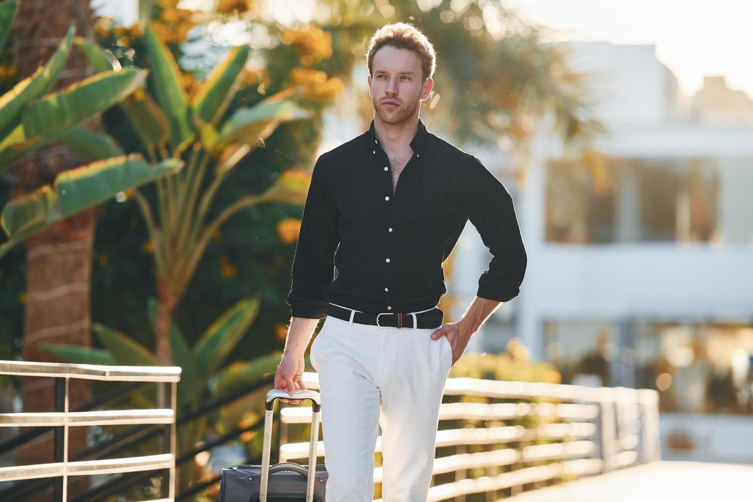 With travel bag. Young man is outdoors at sunny daytime. Concept of vacation photo