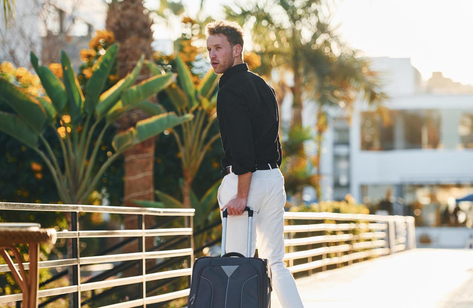 With travel bag. Young man is outdoors at sunny daytime. Concept of vacation photo