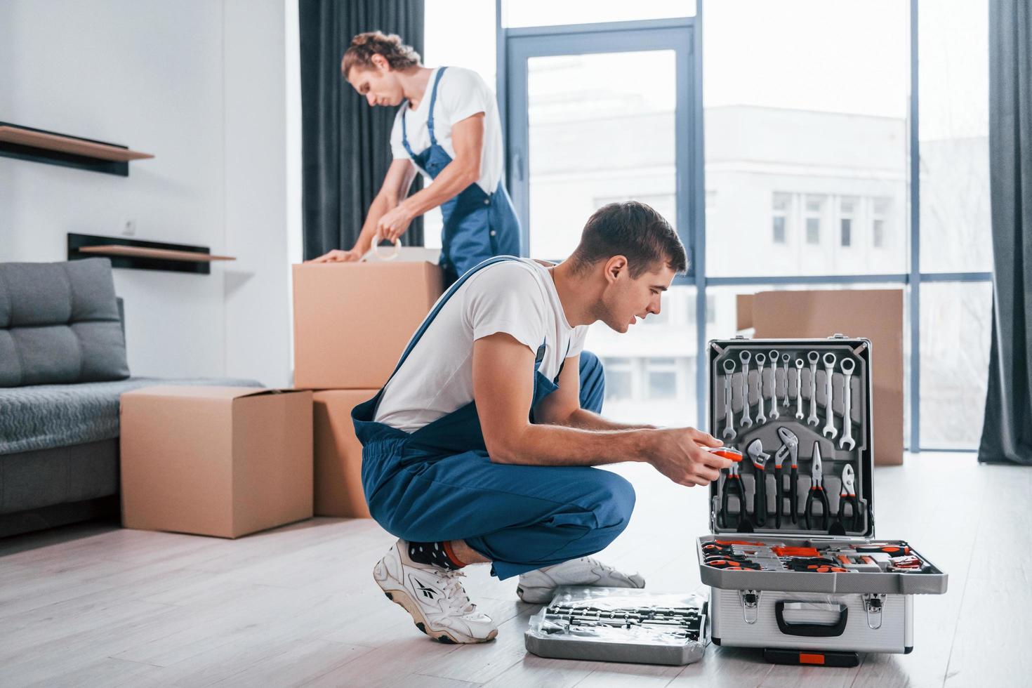 Uses tools from case. Two young movers in blue uniform working indoors in the room photo