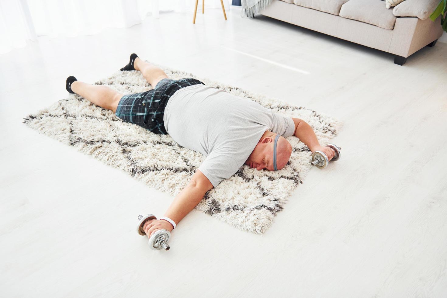 Laying down exhausted. With dumbbells in hands. Funny overweight man in casual clothes is indoors at home photo