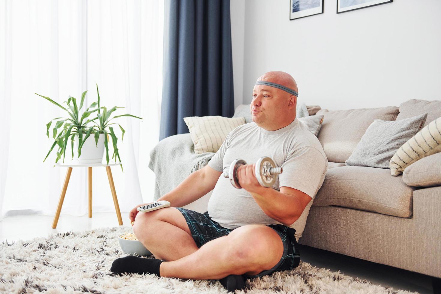 Eats and does exercises. Funny overweight man in casual clothes is indoors at home photo