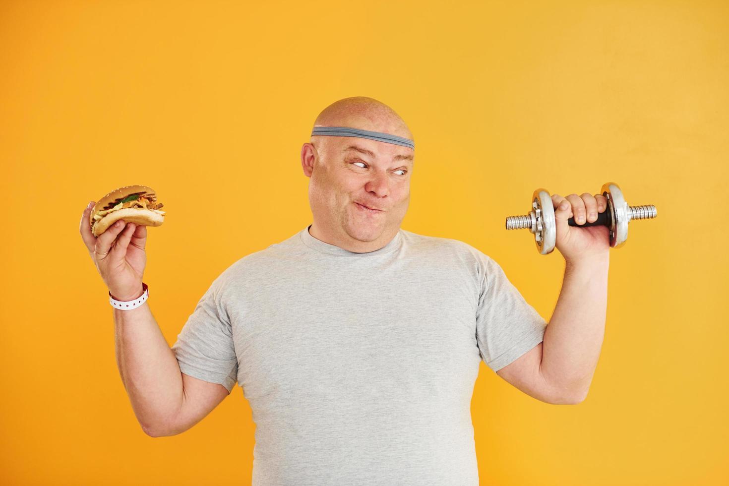 Holds hamburger and dumbbell. Funny overweight man in sportive head tie is against yellow background photo