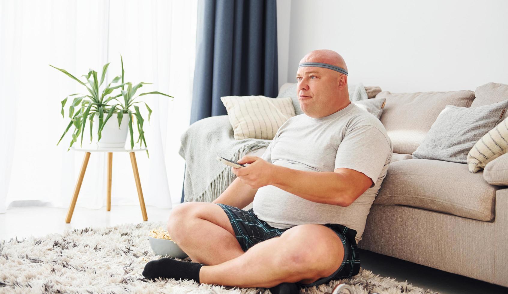 Sits on the sofa with popcorn. Funny overweight man in casual clothes is indoors at home photo