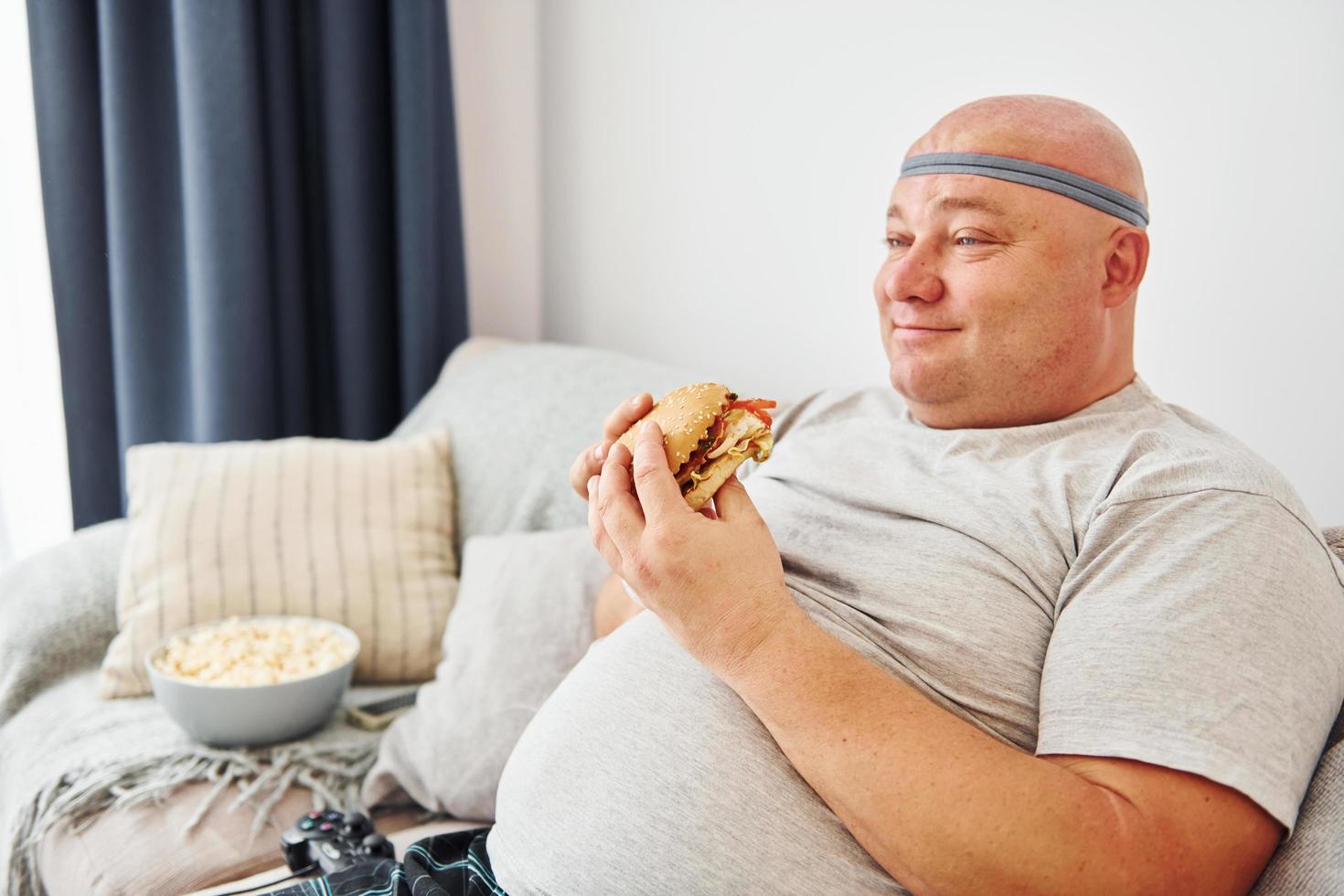 Sits on the sofa with popcorn and hamburger. Funny overweight man in casual clothes is indoors at home photo