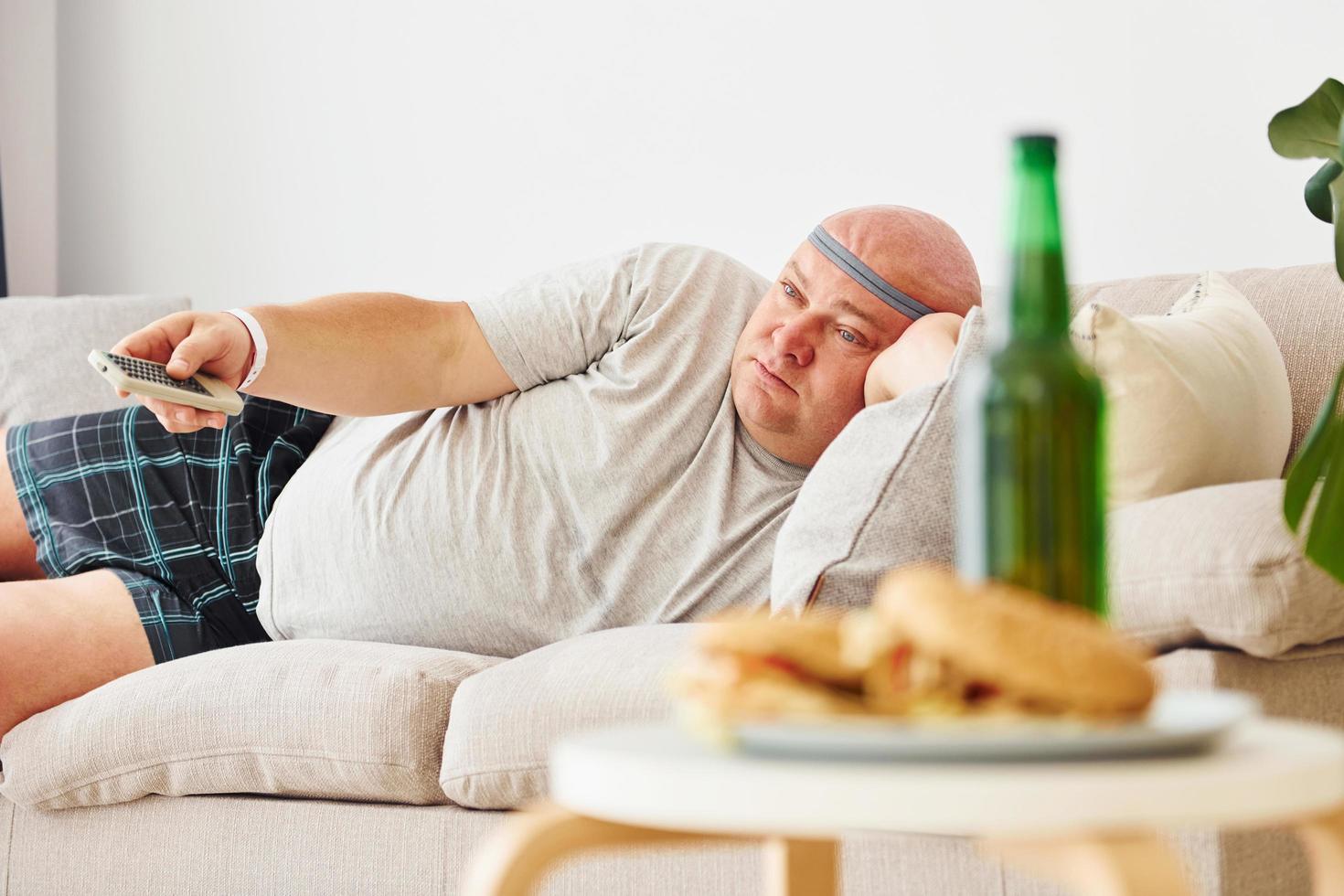 hamburguesas y cerveza. hombre acostado en el sofá. un hombre gracioso con sobrepeso vestido con ropa informal está en el interior de su casa foto