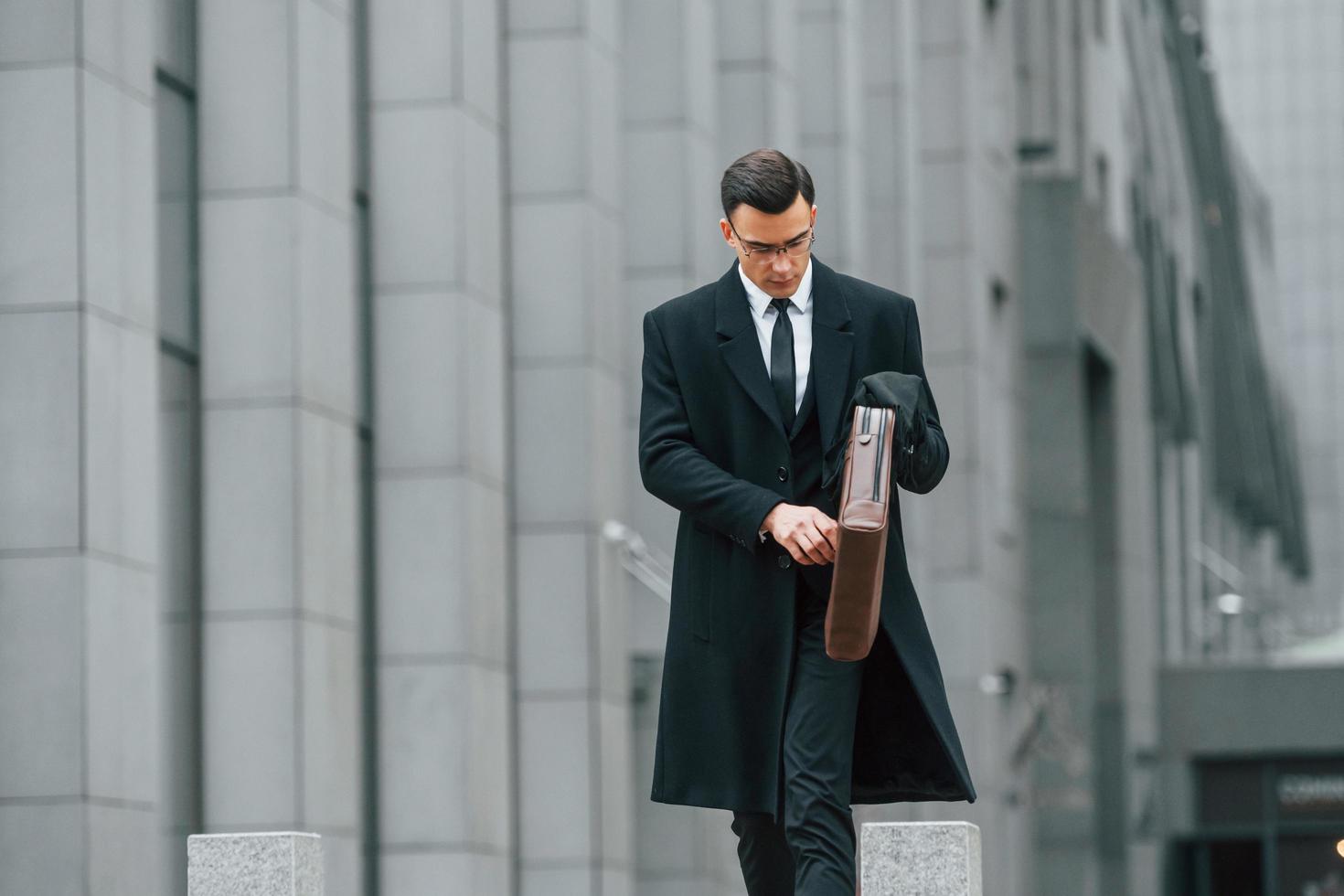 Holding brown bag in hands. Businessman in black suit and tie is outdoors in the city photo