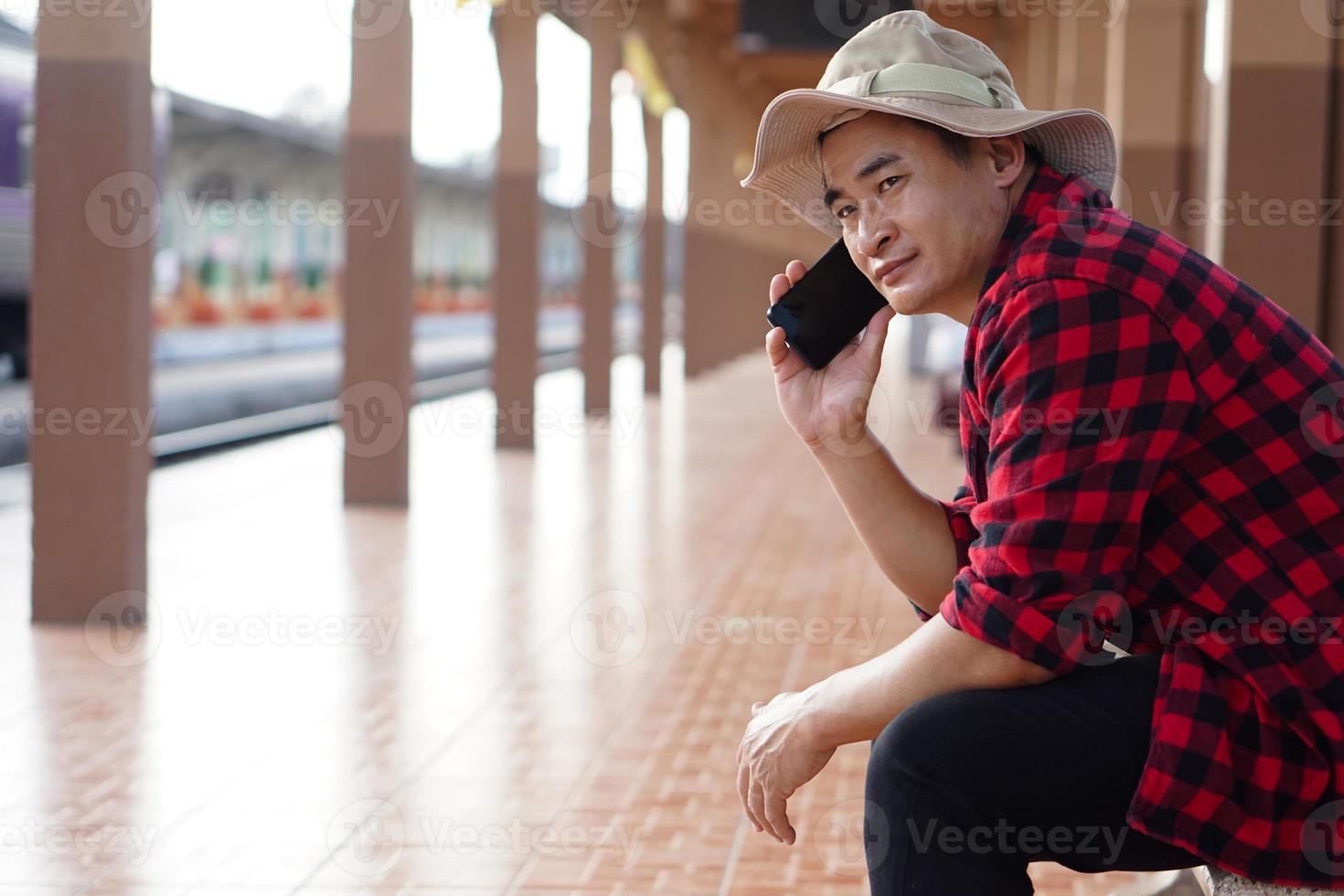 Asian man traveler is waiting someone at platform of train station, holds mobilephone to talk on the phone. Concept, travel alone, transportation, communication or connection. photo