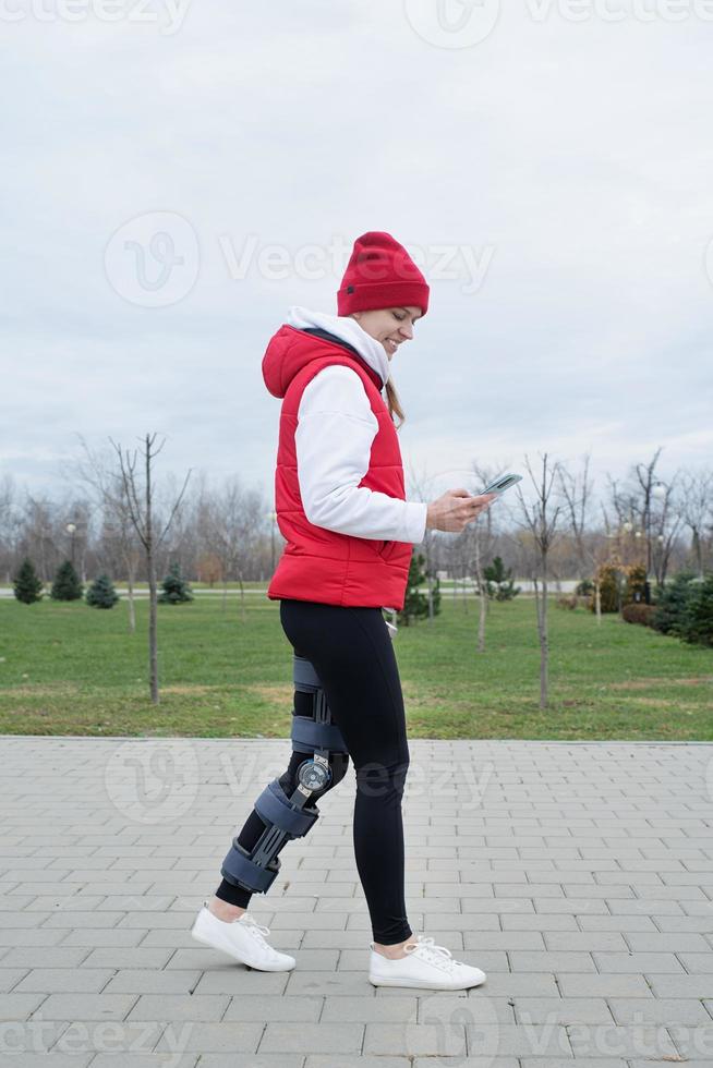 Woman wearing knee brace or orthosis after leg surgery walking in the park using smartphone photo