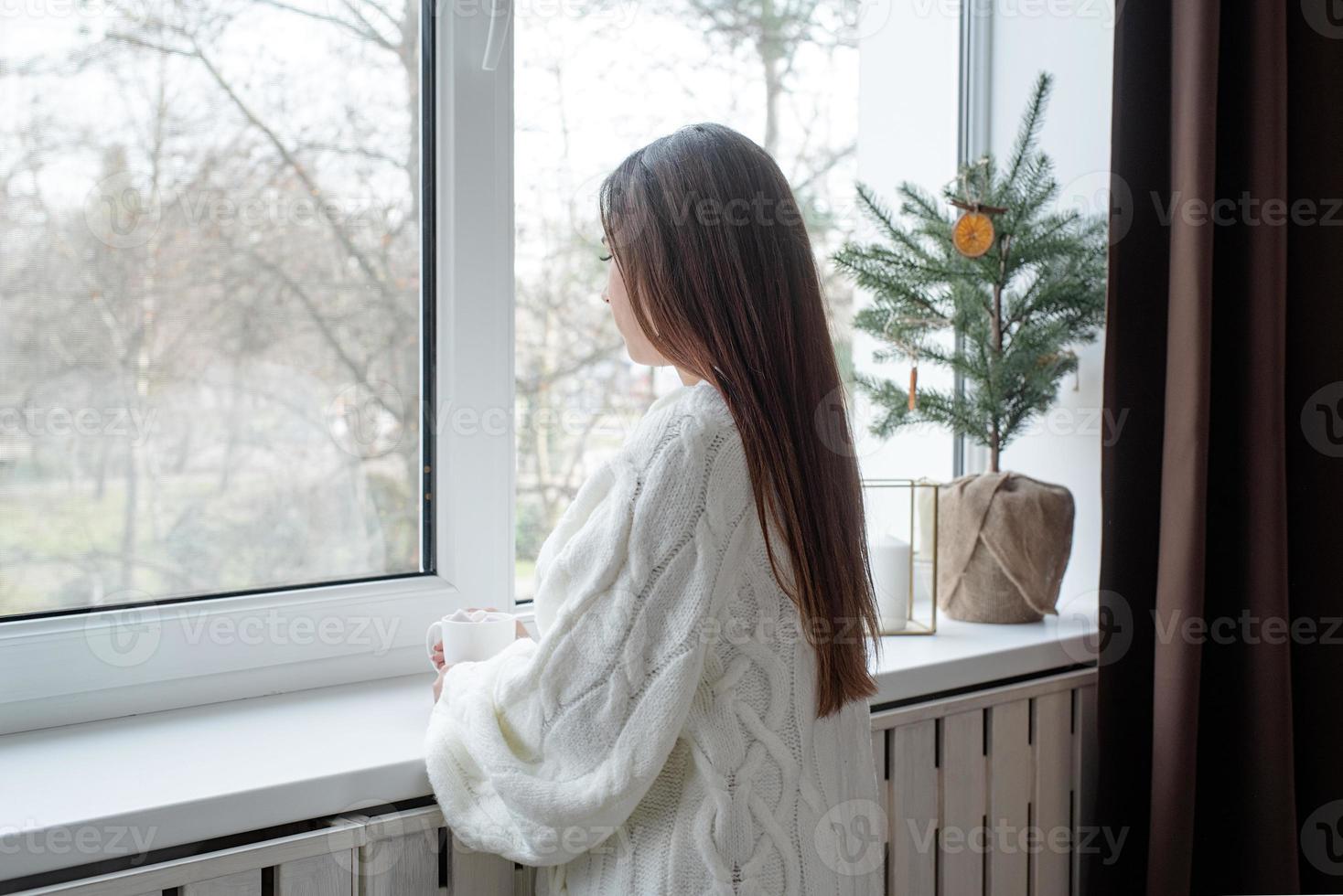 Woman in warm white winter sweater standing next to the window at home at christmas eve holding cup with marshmallows, fir tree behind photo